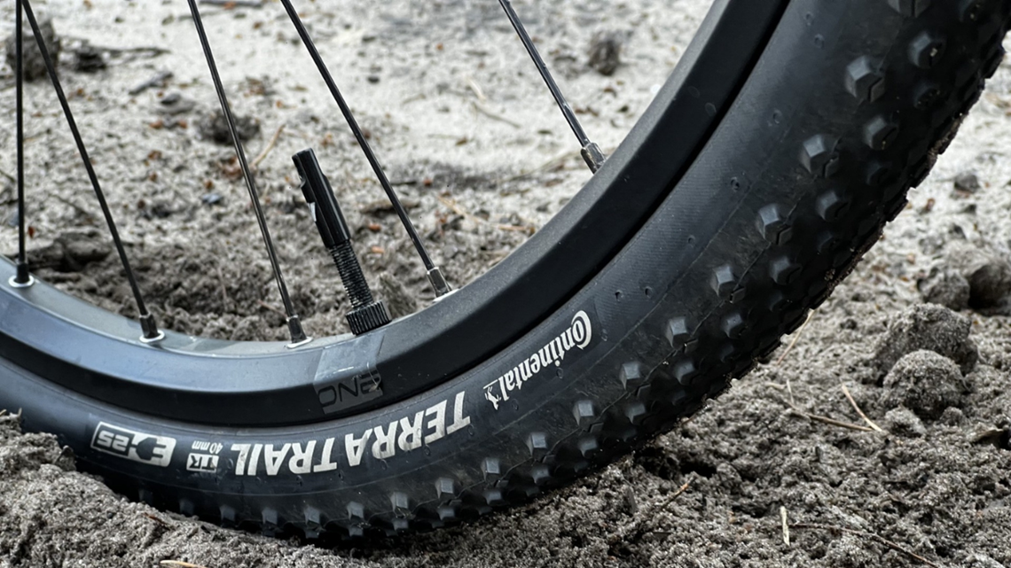 Zbliżenie na czarną oponę rowerową Continental Terra Trail zagłębioną w sypkim, szarym piasku. Widoczny bieżnik z klockami oraz napis 
